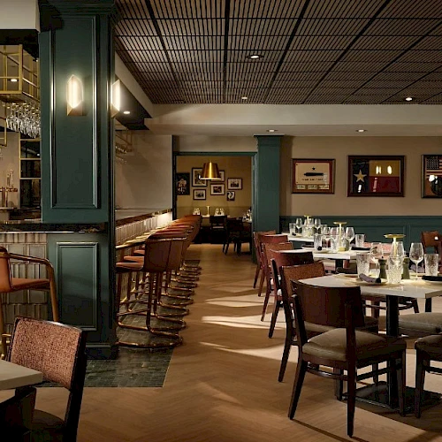 A stylish, dimly lit restaurant with green walls, wooden chairs, round and square tables set with glassware, and a bar area to the left, ready for guests.