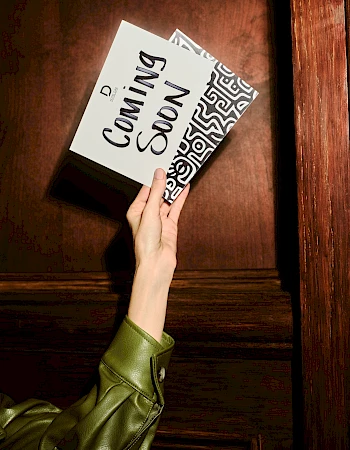 A hand pulls a book titled “Coming Soon” from a wooden shelf, with bold black text and a patterned cover, in a dramatic close-up.