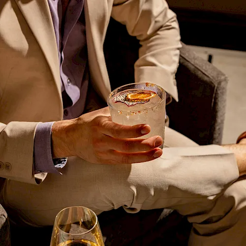 Two people in suits enjoying drinks; one holds a glass with a light-colored cocktail, the other has a whiskey glass, in a social setting.