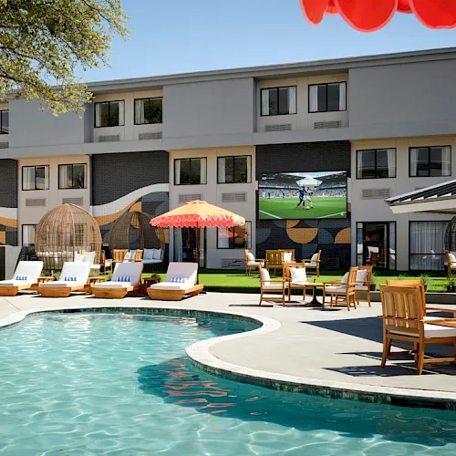 A sunny hotel pool area with lounge chairs, umbrellas, and a European-style courtyard; guests relax while a flat-screen TV shows a sports game.