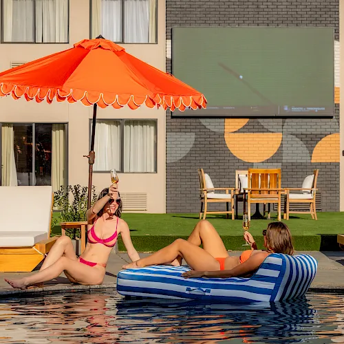 Two women lounge poolside on a sunny day under an orange umbrella, sipping champagne in bikinis while others relax on sunbeds.