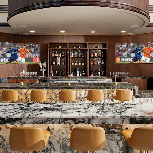 A sleek bar area with marble countertops, row of brown bar stools, backlit shelves, and two TV screens showing sports. End the sentence with a period.