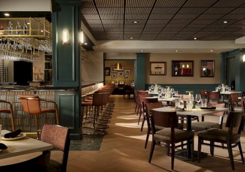 A stylish, dimly lit restaurant with green walls, wooden chairs, round and square tables set with glassware, and a bar area to the left, ready for guests.