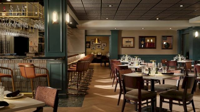 A stylish, dimly lit restaurant with green walls, wooden chairs, round and square tables set with glassware, and a bar area to the left, ready for guests.