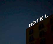 A lit HOTEL sign crowns a brick building at dusk, glowing against a dark blue sky.