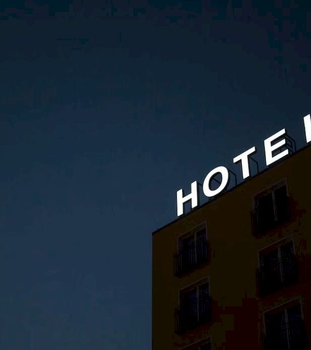 A lit HOTEL sign crowns a brick building at dusk, glowing against a dark blue sky.