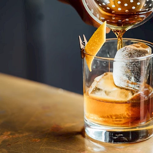 A whiskey cocktail with a large ice cube being poured into a rocks glass, garnished with an orange twist and a small skewer.