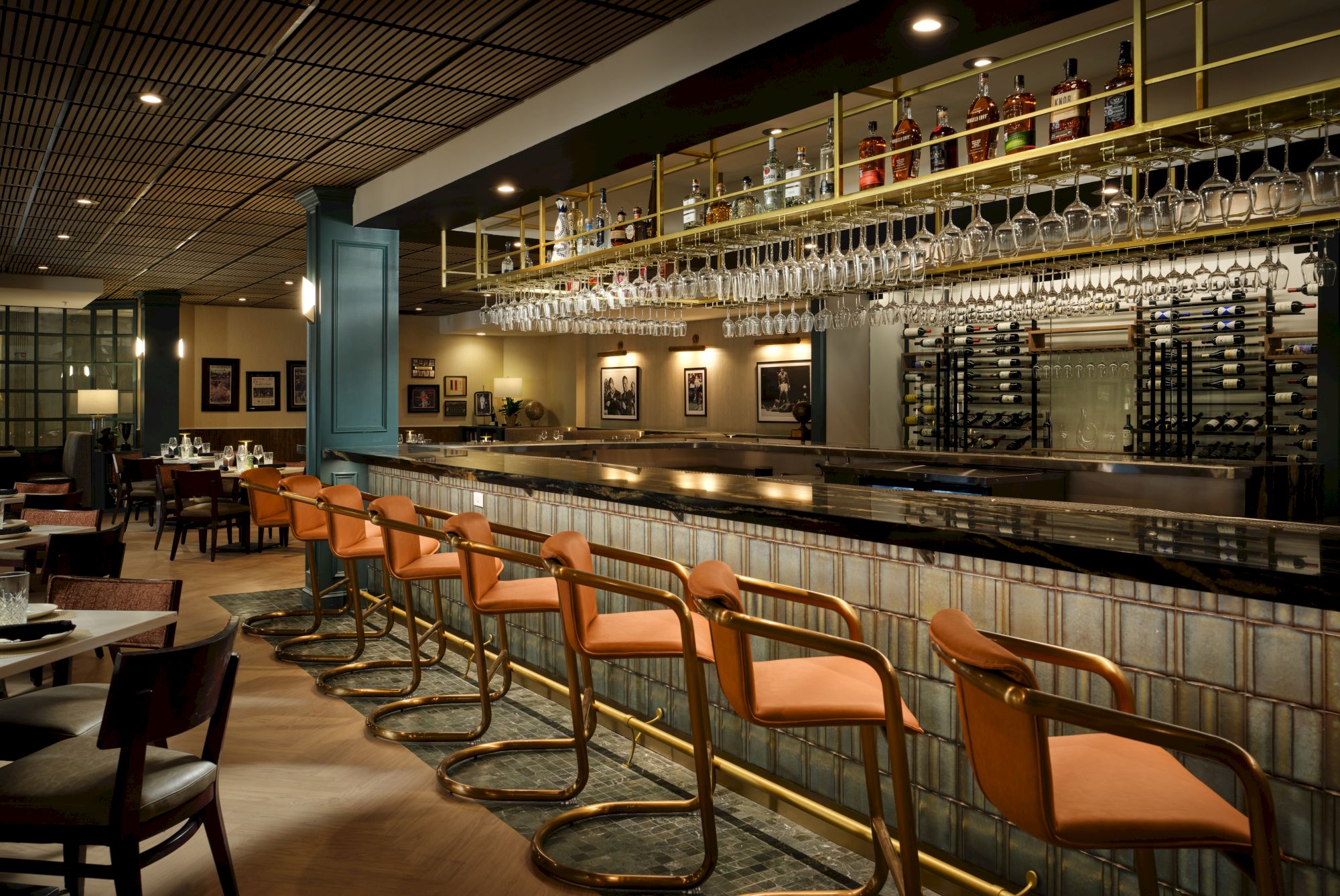 A sleek bar with orange stools, a long tiled counter, hanging glassware, dim seating, and framed art on the walls, cozy and stylish.