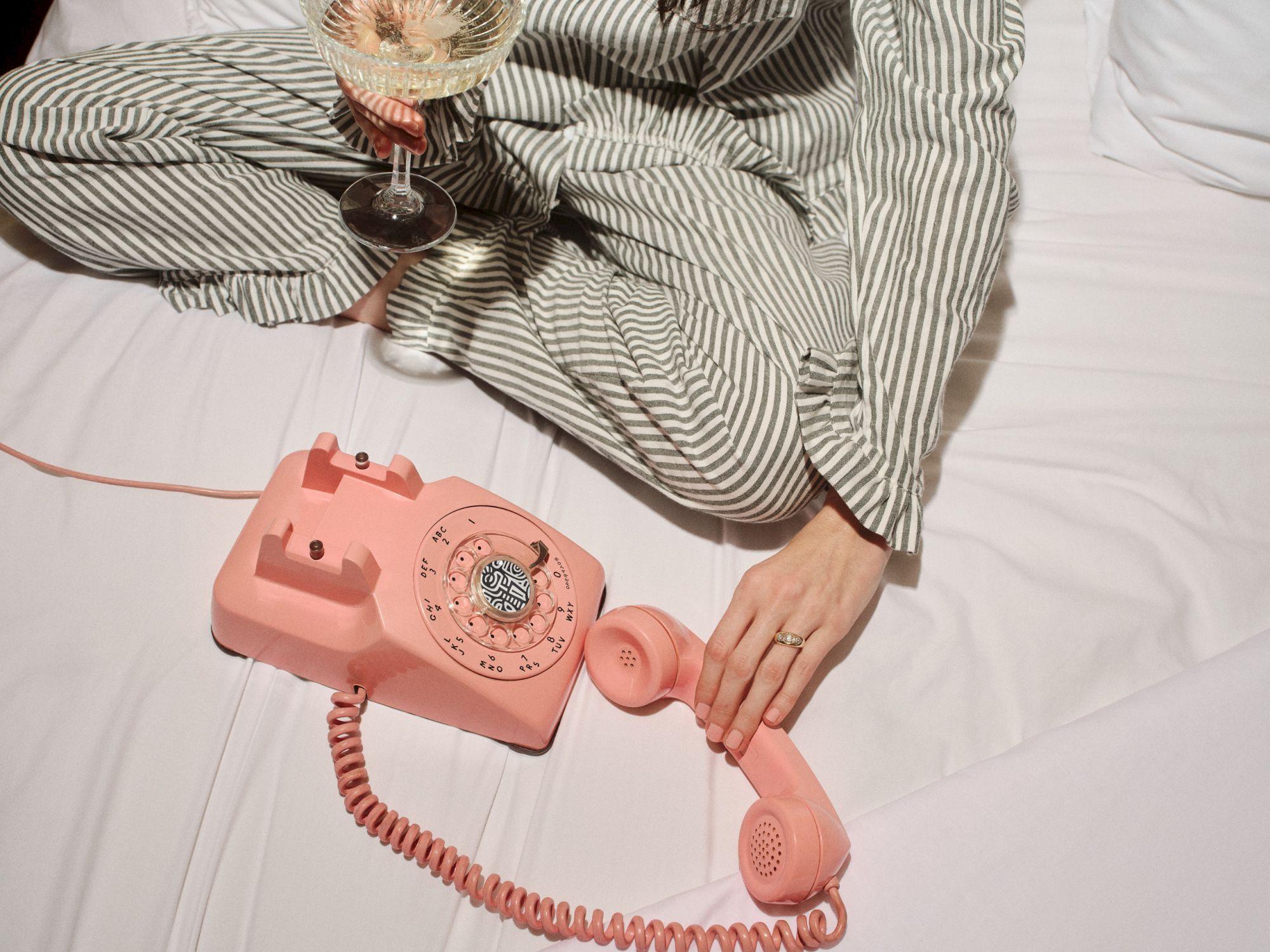 A person in striped pajamas sits on a bed, holding a pink rotary telephone and a glass of wine, with a hand resting on the phone.