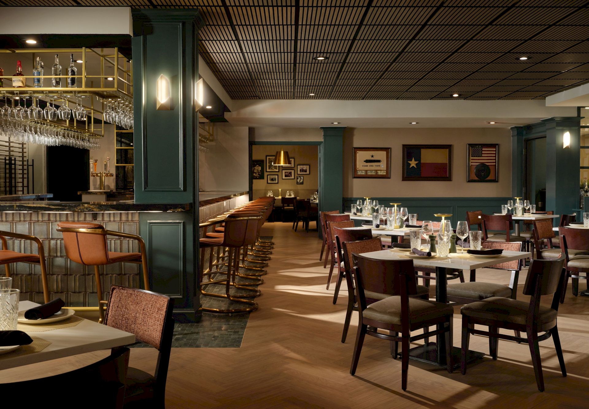 A stylish, well-lit restaurant with wooden tables and chairs, a bar area on the left, framed art on green walls, and cozy seating across the room.