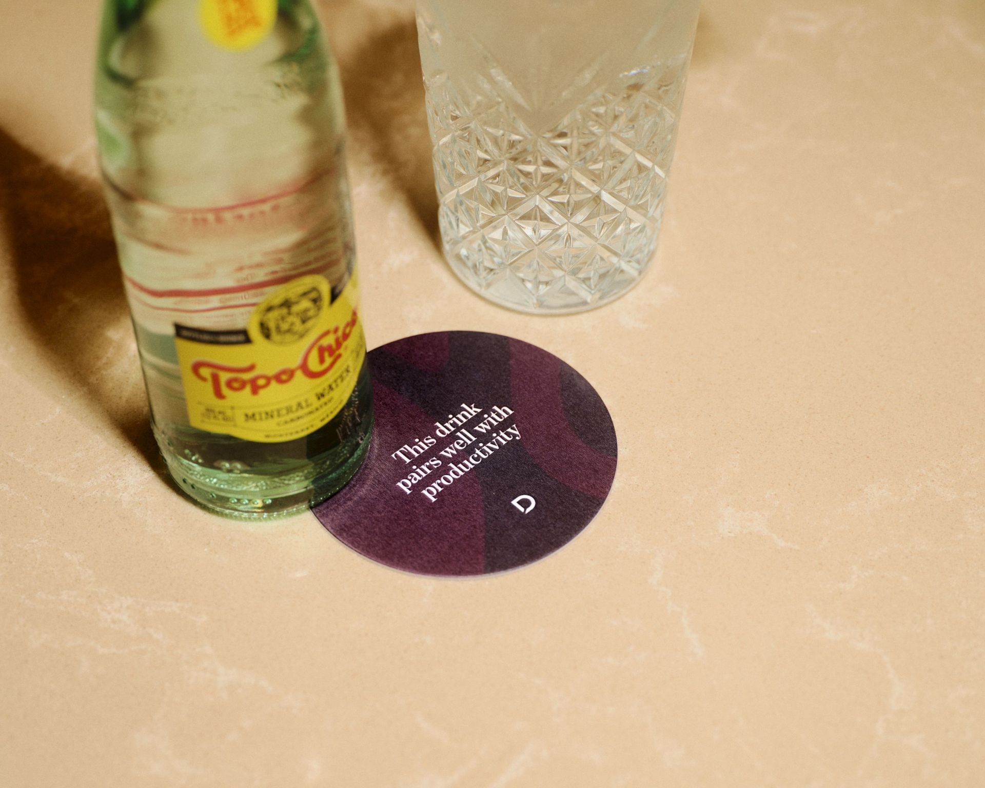 A small bottle of Topo Chico-style soda (green-tinted bottle) sits next to a crystal glass on a table, with a purple circular name tag or coaster nearby.