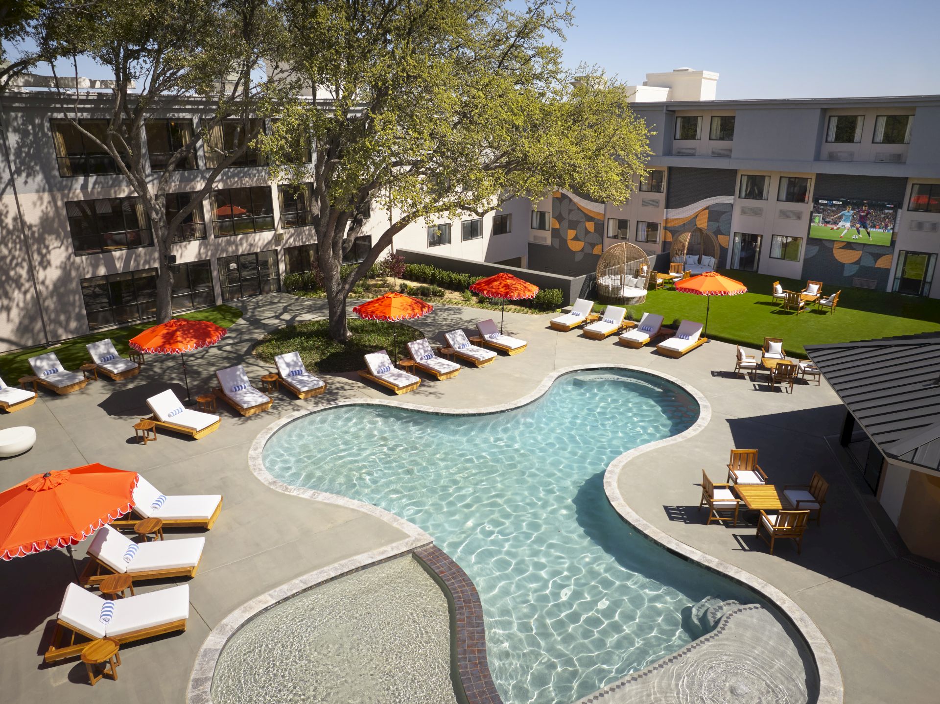 A hotel-style pool area with a curvy, shallow pool, sun loungers, orange umbrellas, shaded seating, and surrounding low-rise buildings.