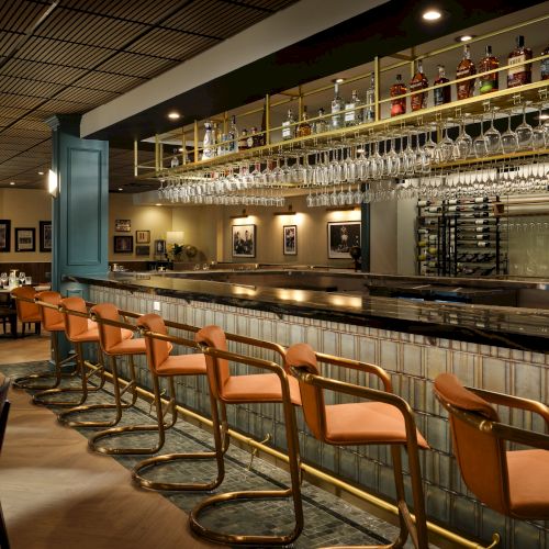 A stylish bar with a long tiled counter, row of orange stools, hanging glassware, and dim dining area in the background, inviting for cocktails tonight.
