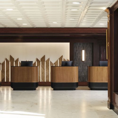 A grand hotel lobby with a long reception desk, warm lighting, and elegant marble floors, framed by artful walls and decorative plants.