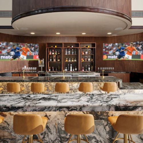 A stylish bar with a marble counter, golden stools, backlit shelves, and two TVs showing a sports program, creating a luxe lounge vibe.