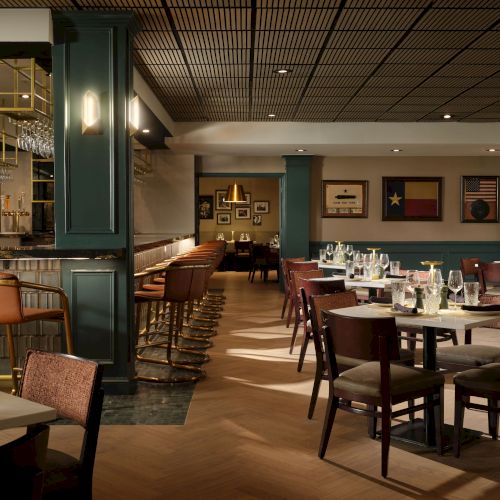 A stylish restaurant interior with wooden floors, green paneling, multiple round and rectangular tables set for dining, wine glasses, and a bar area in the background.