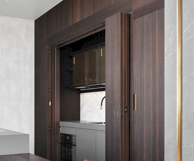 The image shows a modern kitchen with a wood finish, featuring hidden appliances behind pocket doors and adjacent to a dining area.