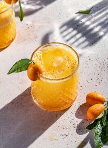 A glass of orange cocktail with ice, garnished with a small citrus fruit and a leaf, sits on a sunlit surface next to some kumquats.