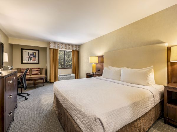 A tidy hotel room with a king bed, two nightstands, lamps, a desk, chair, dresser, and a small window with curtains. Always ends the sentence.