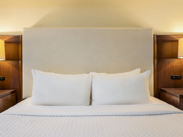 A neatly made hotel bed with two white pillows, padded headboard, and warm bedside lamps on wooden nightstands. classic, cozy, inviting.