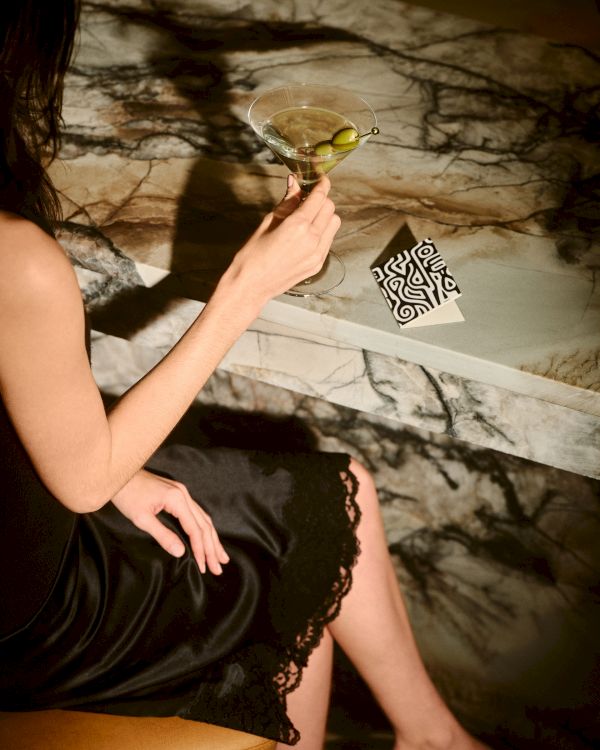 A woman in a black dress sits with a cocktail, a lime garnish, and a patterned card on a marble surface, partial view of legs visible.