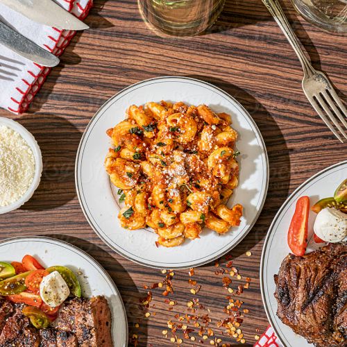 The image shows a meal set on a wooden table: pasta with red sauce, steak, salad, grated cheese, cutlery, and glasses.