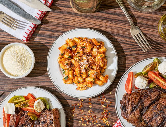 The image shows a meal set on a wooden table: pasta with red sauce, steak, salad, grated cheese, cutlery, and glasses.