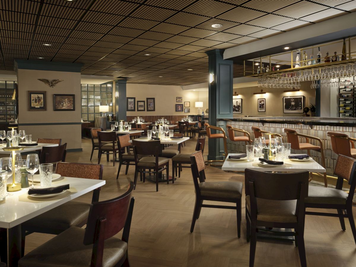 A stylish, dimly lit restaurant with neatly set tables, modern chairs, a bar area, and framed art on the walls, ready for guests.