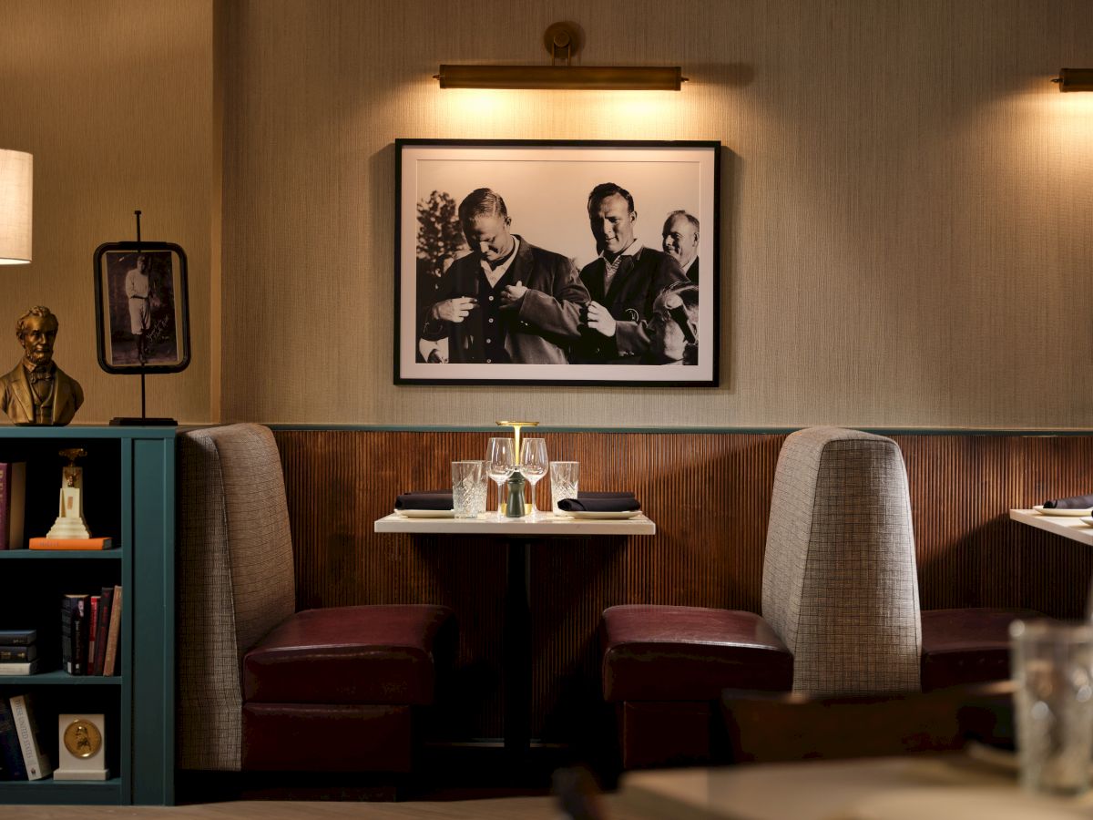 A cozy restaurant booth with two tables and red seats, framed black-and-white photos on the wall, warm lighting, and a small bookshelf nearby.