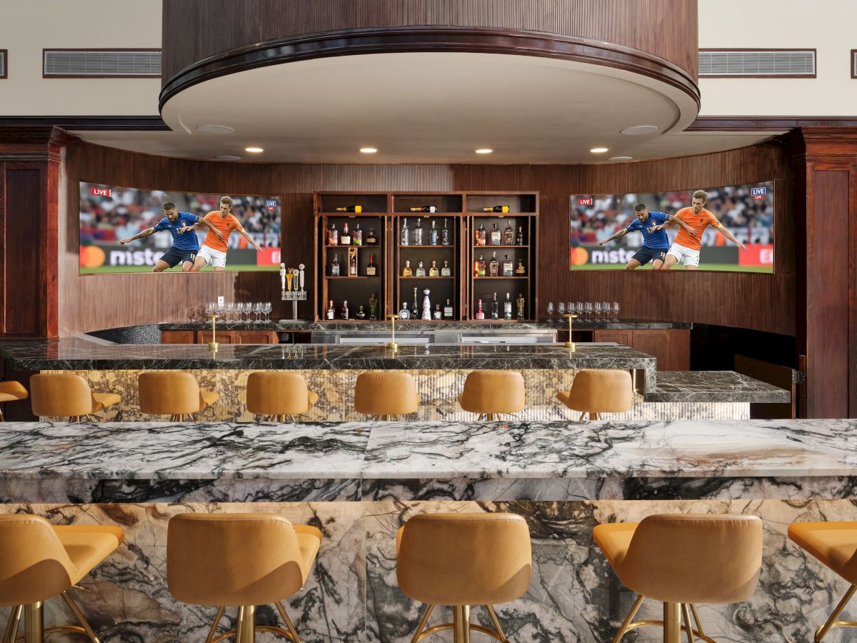 A stylish hotel bar with a marble counter, warm tan stools, and two TVs behind the shelves showing a sports broadcast.