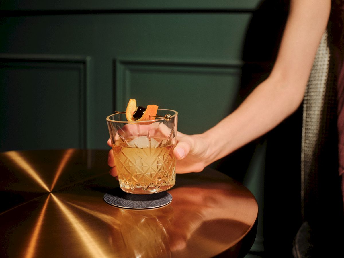 A glass tumbler with a diamond-cut pattern sits on a copper bar, garnished with a twist of orange peels, held by a person.