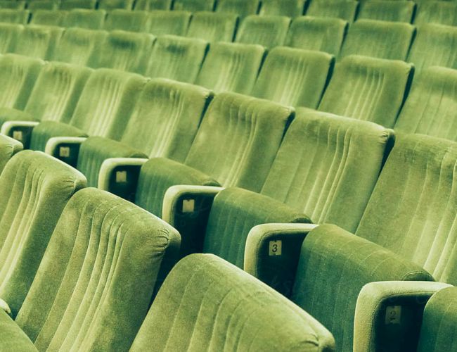 A row of empty green theater or stadium seats, evenly spaced and slightly worn, creating a repeating pattern and a quiet, overlooked space.