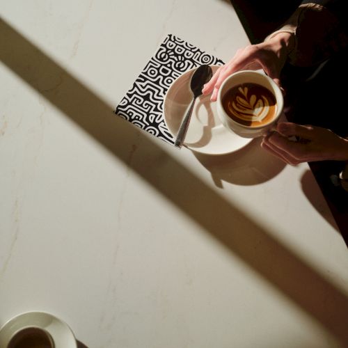 A person pours latte art into a cup from above, with sunlight casting long shadows on a white table and a patterned coaster nearby.
