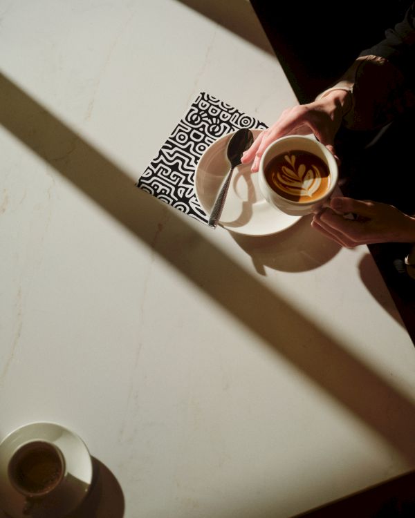 A person pours latte art into a cup from above, with sunlight casting long shadows on a white table and a patterned coaster nearby.