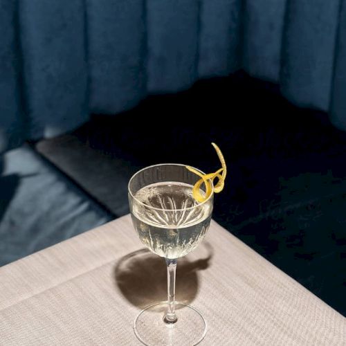 A glass with a light-colored drink and a lemon twist garnish sits on a table with a blue curtain backdrop.