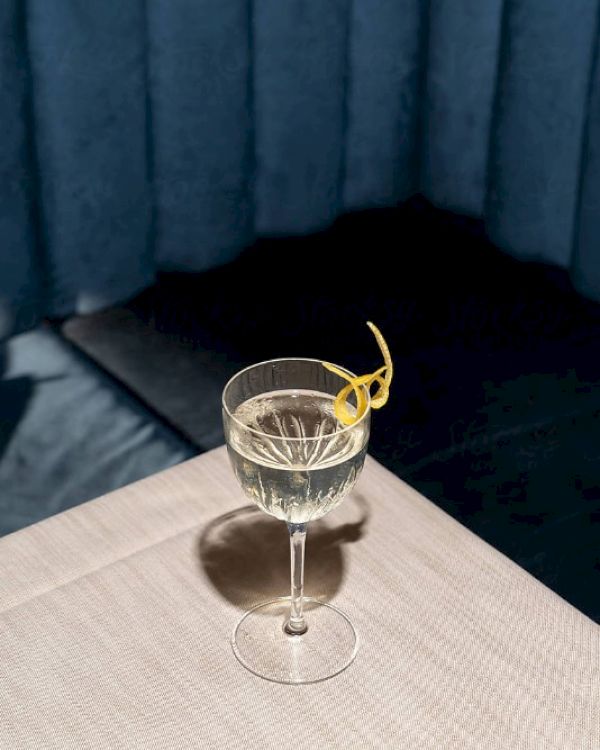 A glass with a light-colored drink and a lemon twist garnish sits on a table with a blue curtain backdrop.