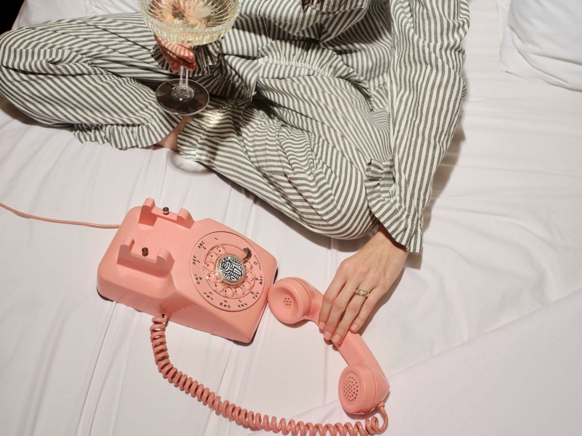 A person in striped pajamas holds a pink vintage rotary desk phone while sipping wine on a bed.