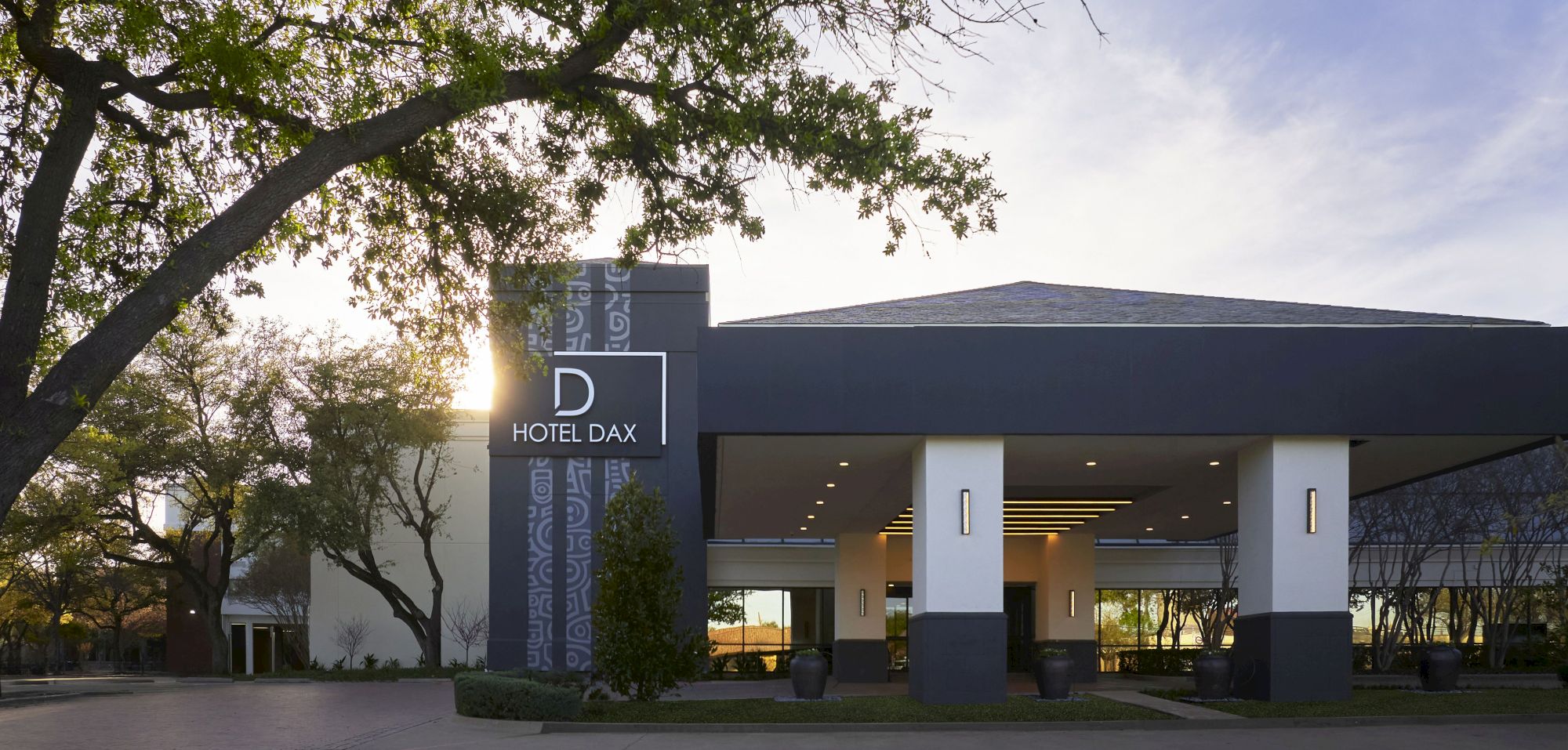 A modern hotel entrance with a covered drive, a tall sign reading &ldquo;HOTEL DAX,&rdquo; trees framing the scene, and a sunny, clear sky.