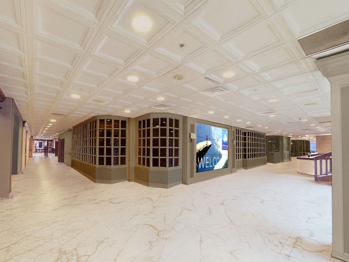 A spacious mall corridor with marble floors, frosted ceiling tiles, built-in display windows, and a large digital ad panel along the curved wall, ending in a far corridor.