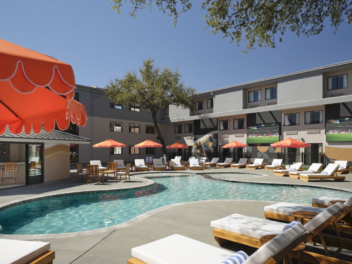 A sunny hotel pool scene with lounge chairs, red umbrellas, and a two-story building in the background.