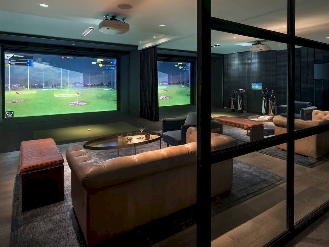 A stylish home theater with multiple screens, a long tan sectional sofa, small ottomans, and a glass-walled layout&mdash;perfect for watching games.