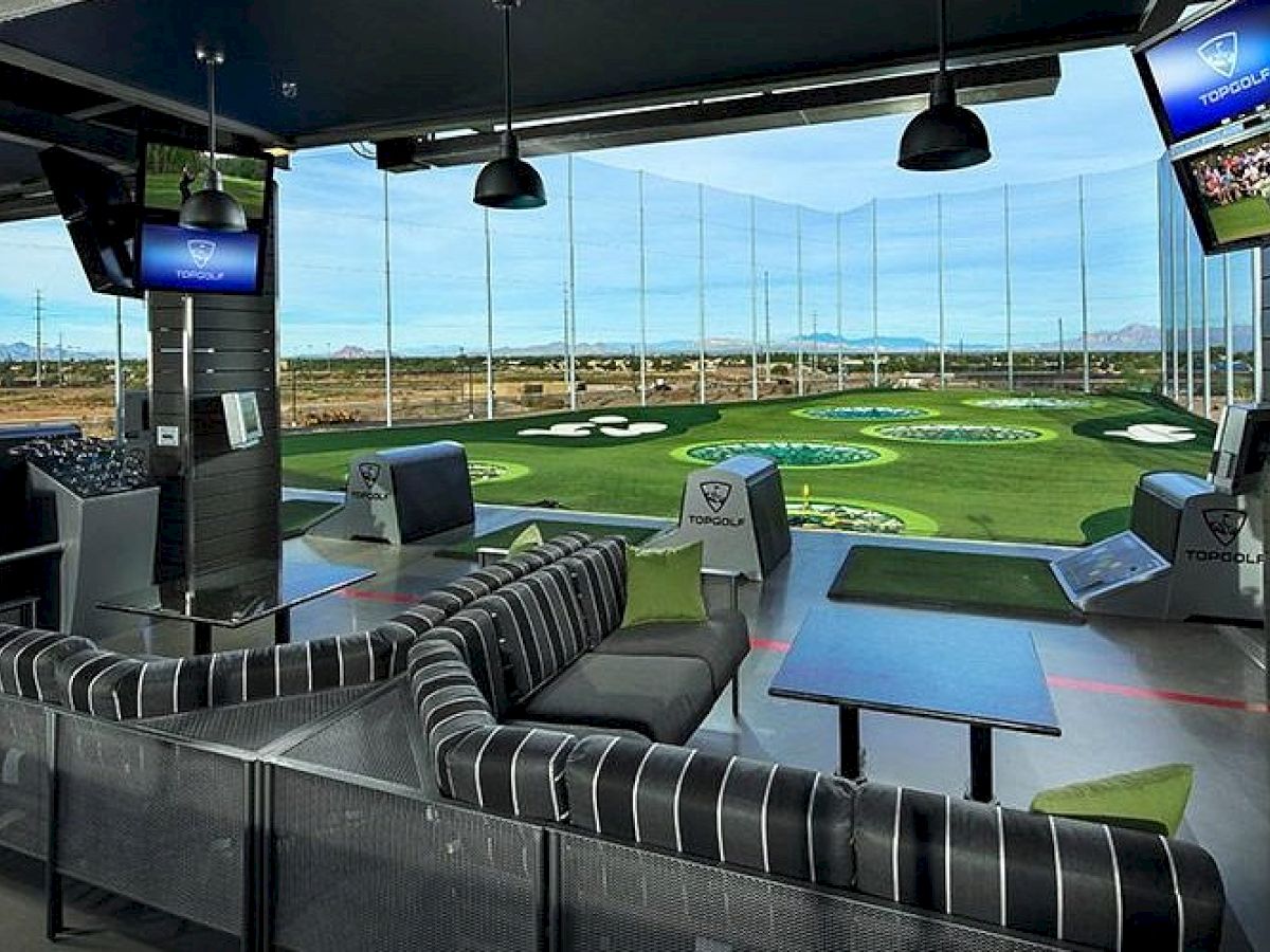 Indoor golf range with seating area, couches, tables, golf simulation screens, and targets outside in open driving range.