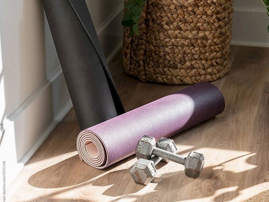 Yoga mat, its carrying tube, a rolled mat, a woven plant pot, and two dumbbells on a sunlit wooden floor.