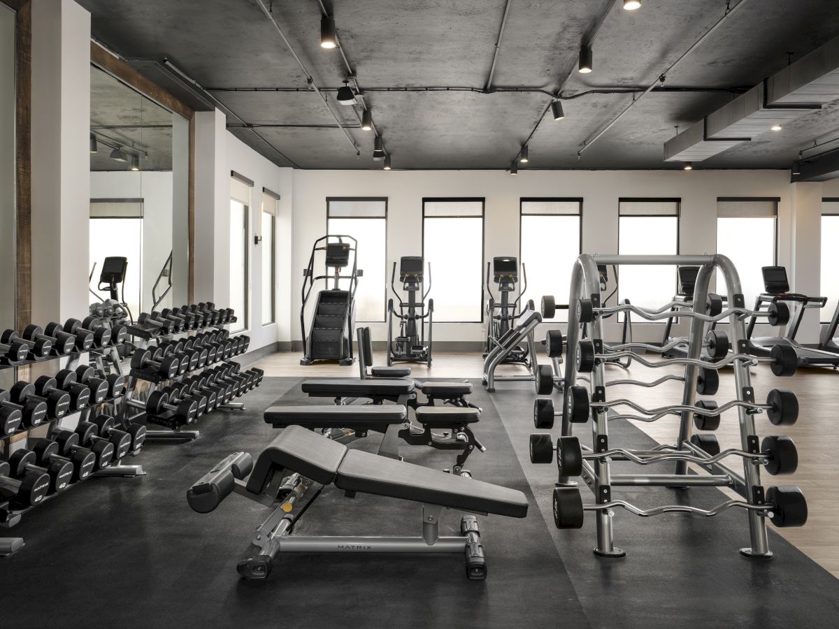 A modern gym with free weights, benches, and a rack of dumbbells, plus cardio machines in the background, all in a spacious industrial setting.