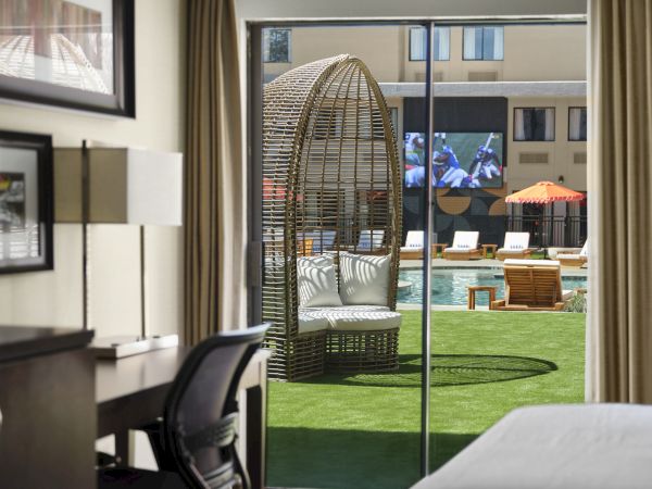A hotel room with a view of a pool area; a wicker dome chair on the lawn, loungers, and a courtyard beyond the glass doors.