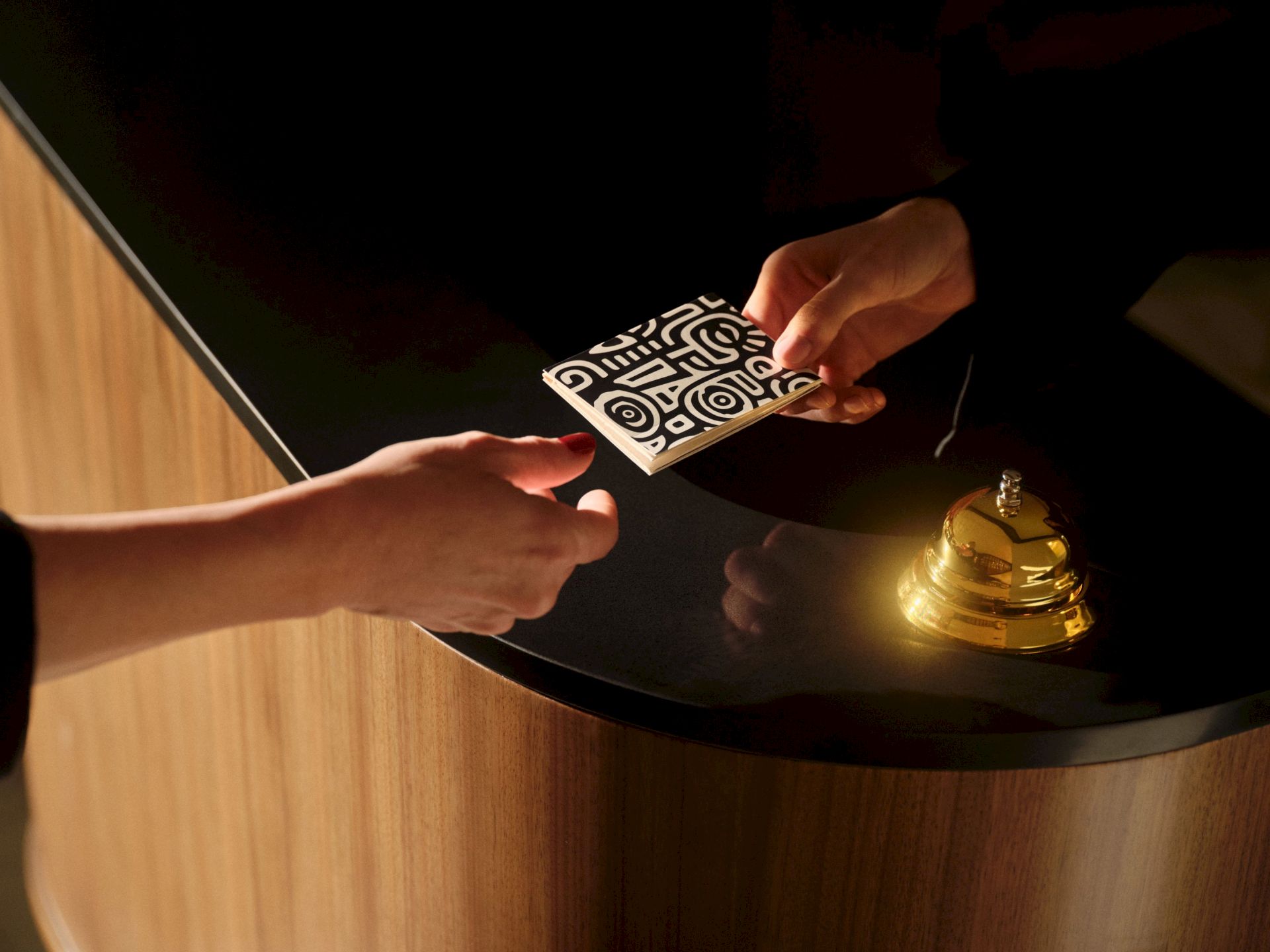 A hand slides a patterned card on a table while another hand reaches, with a warm lamp glowing nearby.