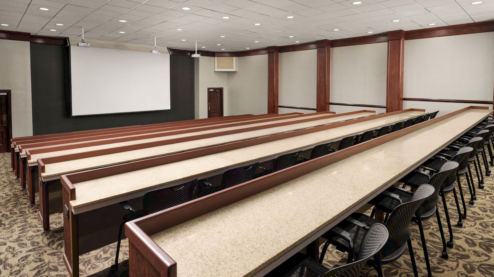 A lecture hall with long classroom tables, black chairs, a projector screen, and recessed ceiling lights.