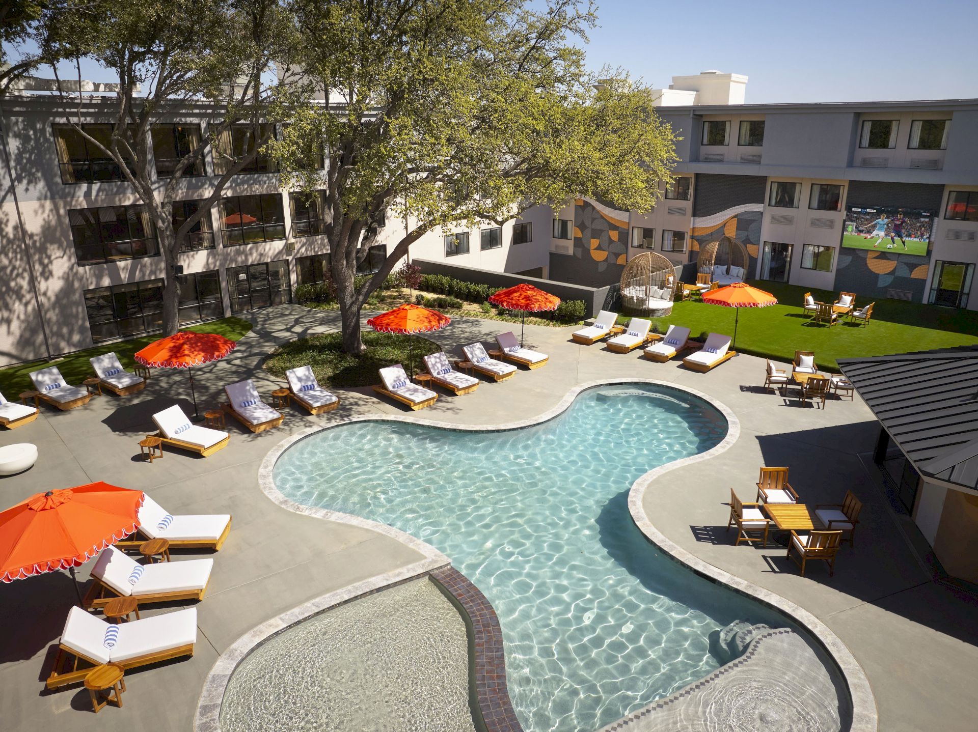 A sunny hotel courtyard with a round pool, lounge chairs under orange umbrellas, trees, and an apartment building with murals.