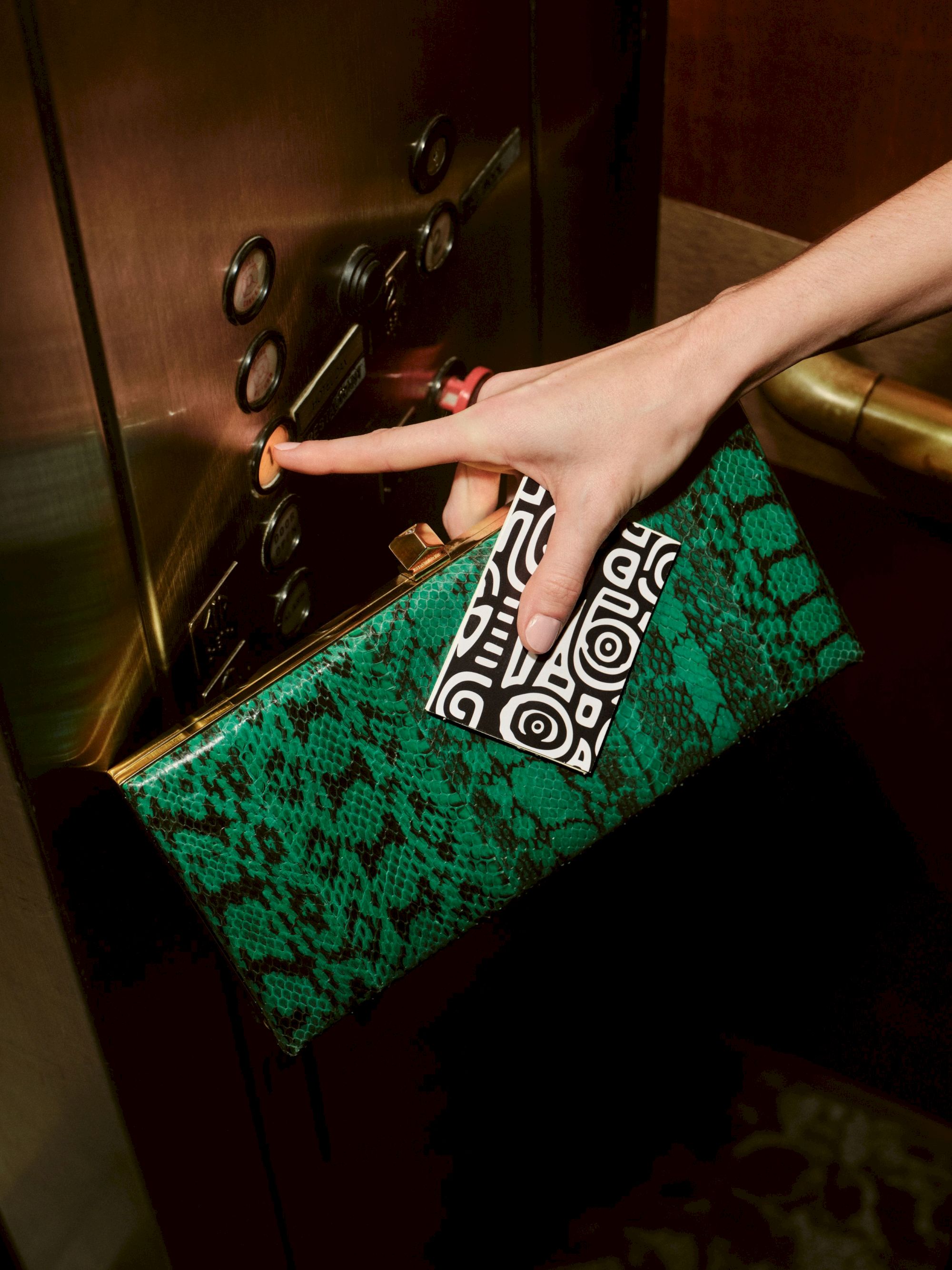 A hand pressing a doorbell or elevator call button, with a patterned black-and-white card near a green patterned bag; action at a panel, ending the sentence.