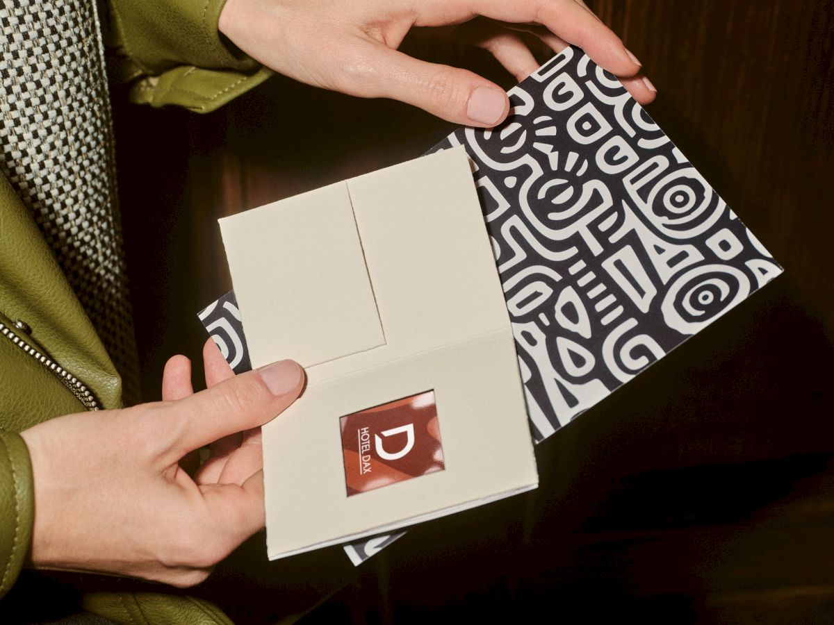 A person opens a small black-and-white patterned box, revealing a white card and a tiny red packaging element inside.